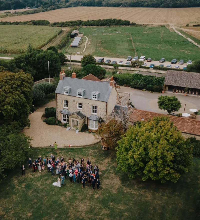 Aerial view of wedding guests gathered on the lawn outside a countryside manor house surrounded by fields and trees.