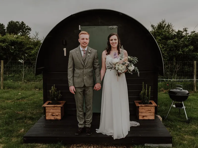 Bride and groom standing hand in hand outside a black shepherd’s hut at East Afton, with the bride holding a bouquet.