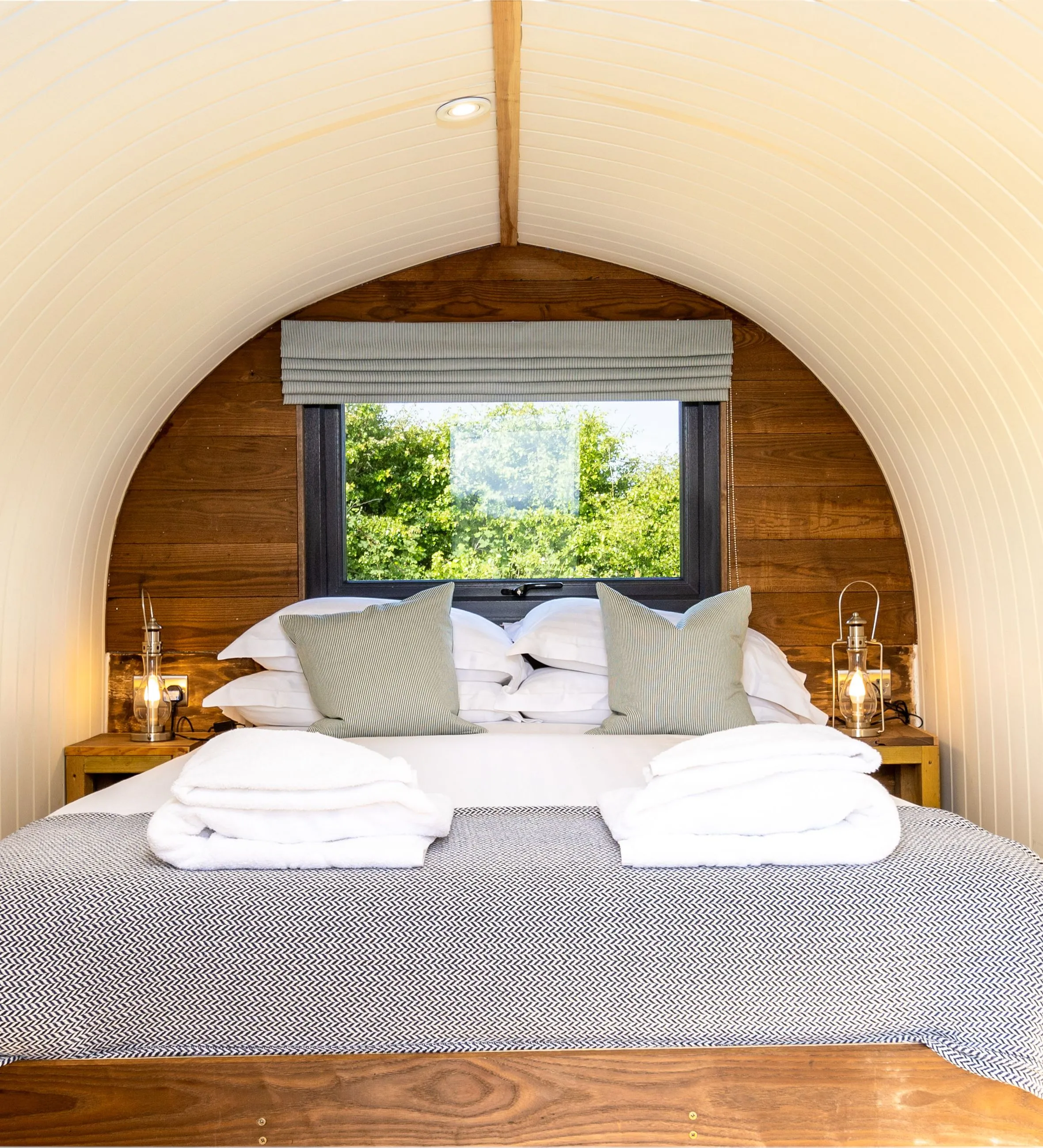 Cosy bedroom inside The Piglets shepherd’s hut with a double bed, bedside lanterns and a window overlooking greenery.