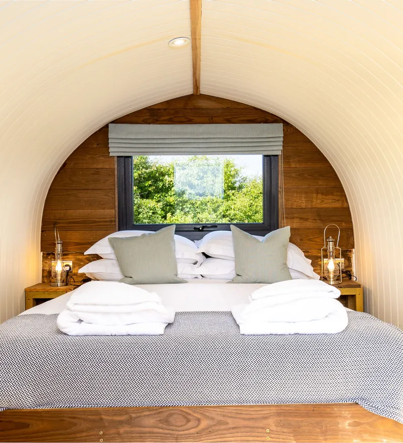 Cosy bedroom inside The Piglets shepherd’s hut with a double bed, bedside lanterns and a window overlooking greenery.