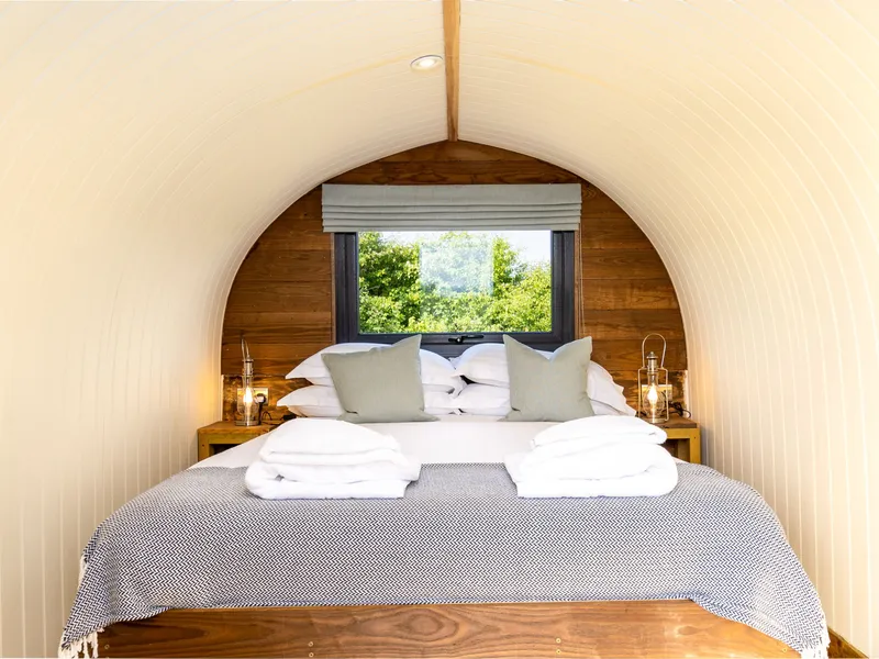 Cosy bedroom inside The Piglets shepherd’s hut with a double bed, bedside lanterns and a window overlooking greenery.