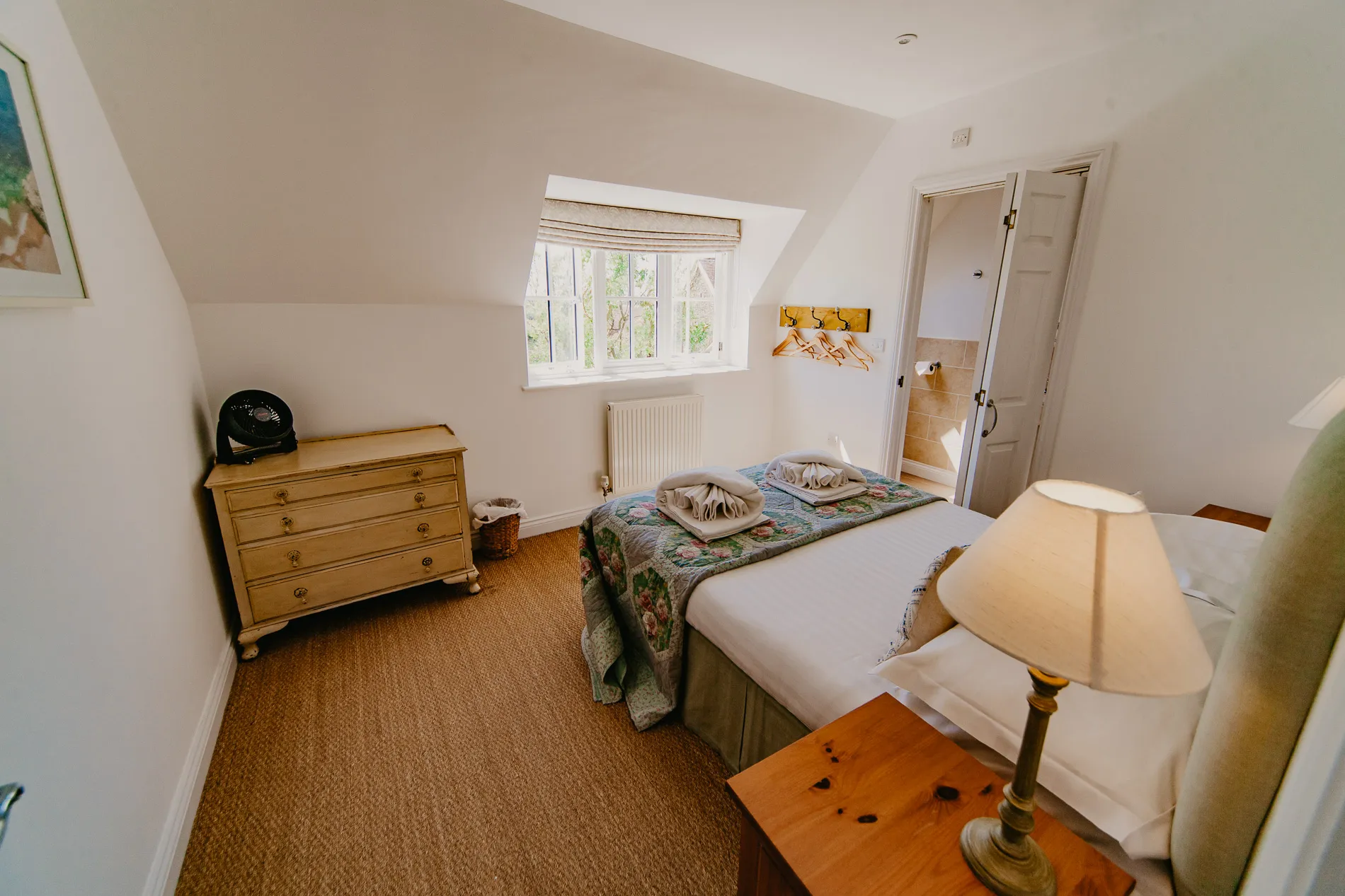 Bright cottage bedroom with double bed, floral quilt, bedside lamp and window, with en-suite door ajar