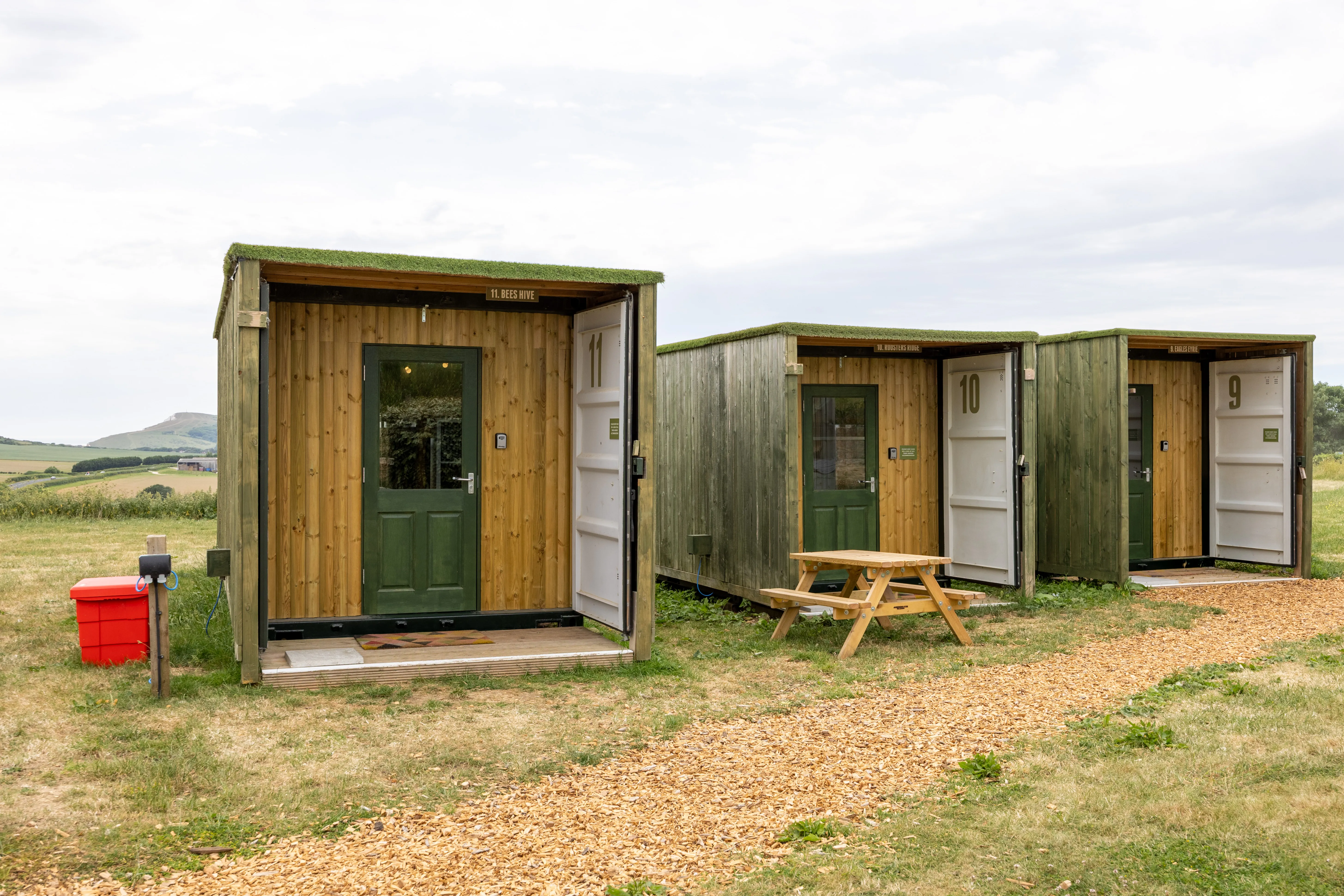 Three countryside glamping cabins at Tapnell Farm with green doors, open fronts and a picnic bench beside a woodchip path.