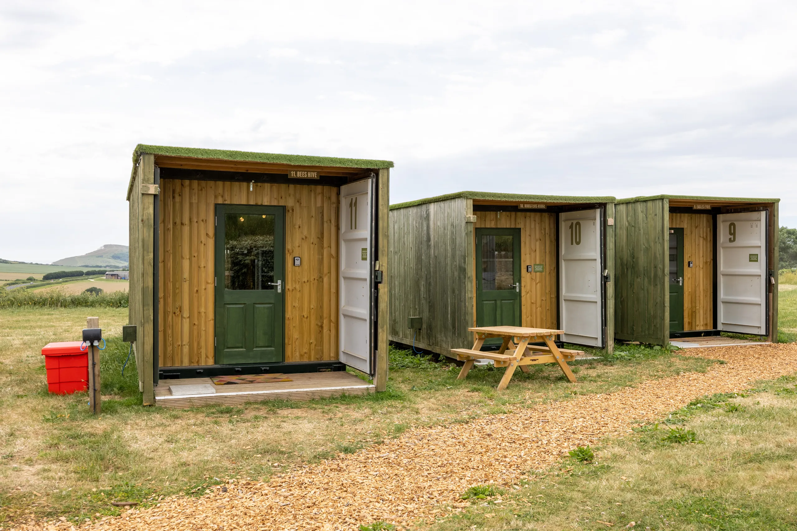 Three countryside glamping cabins at Tapnell Farm with green doors, open fronts and a picnic bench beside a woodchip path.