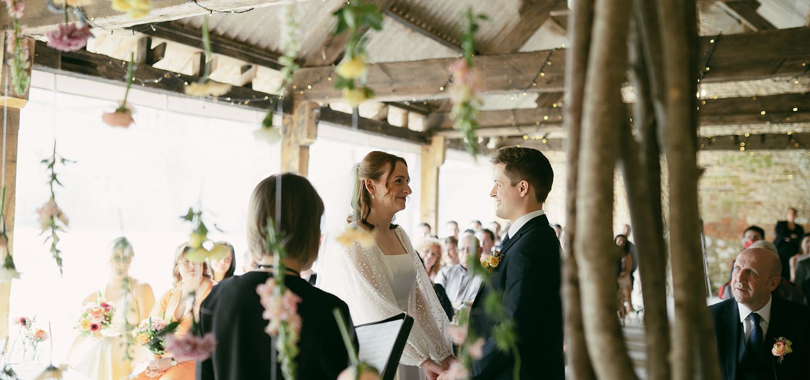 Bride and groom standing hand in hand during their wedding ceremony at East Afton, framed by hanging flowers and rustic wooden beams.