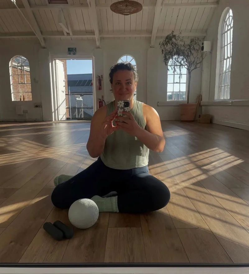 Woman seated cross-legged in a bright studio taking a mirror selfie, with a massage ball and hand weights on the floor in front of her.