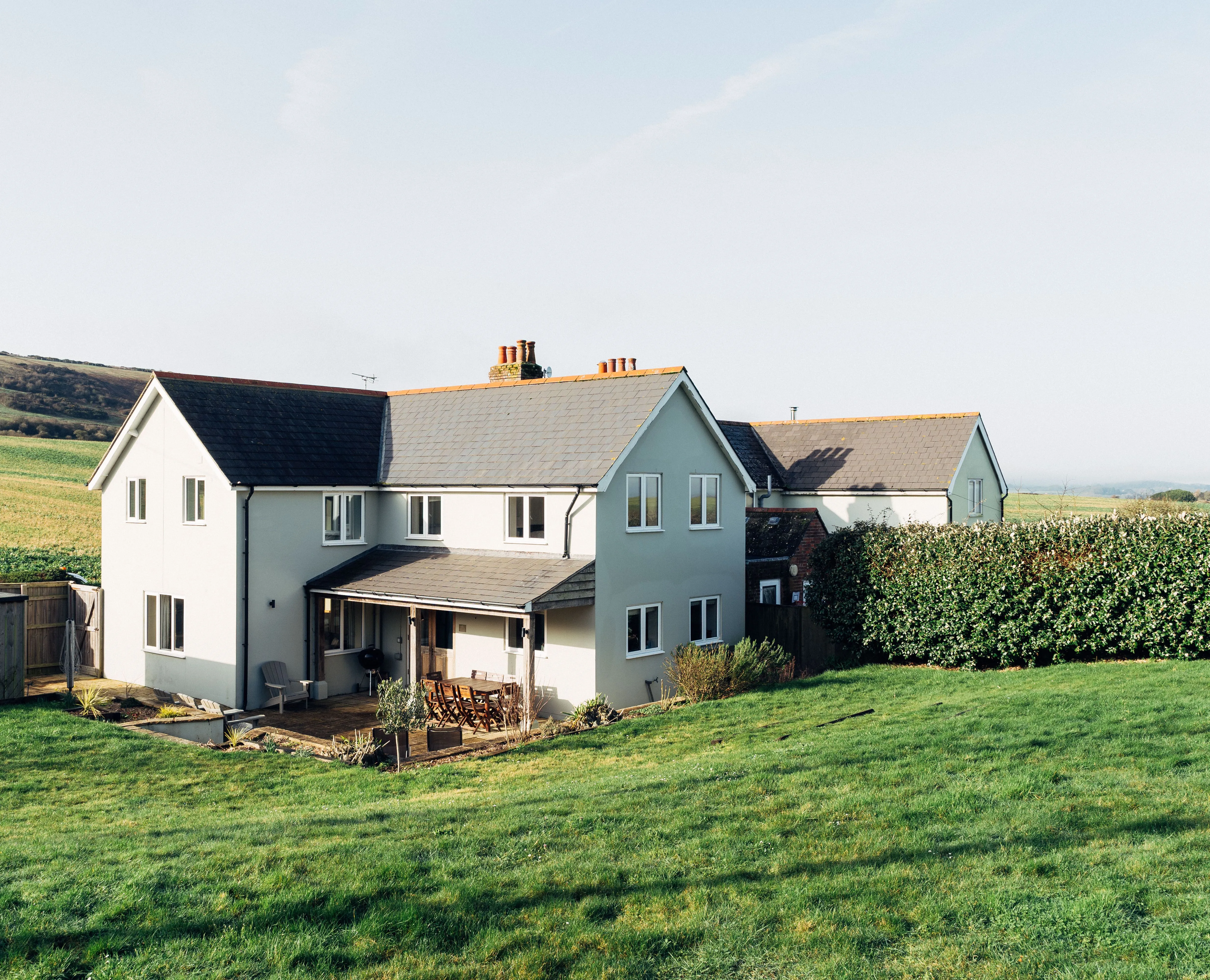 Modern countryside house with a patio and lawn, set against rolling green hills under a clear sky.