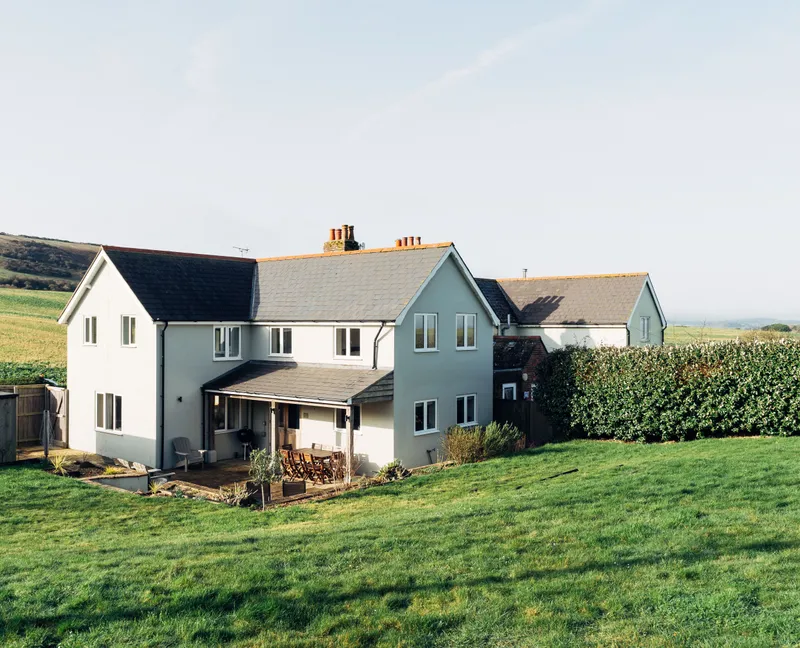 Modern countryside house with a patio and lawn, set against rolling green hills under a clear sky.