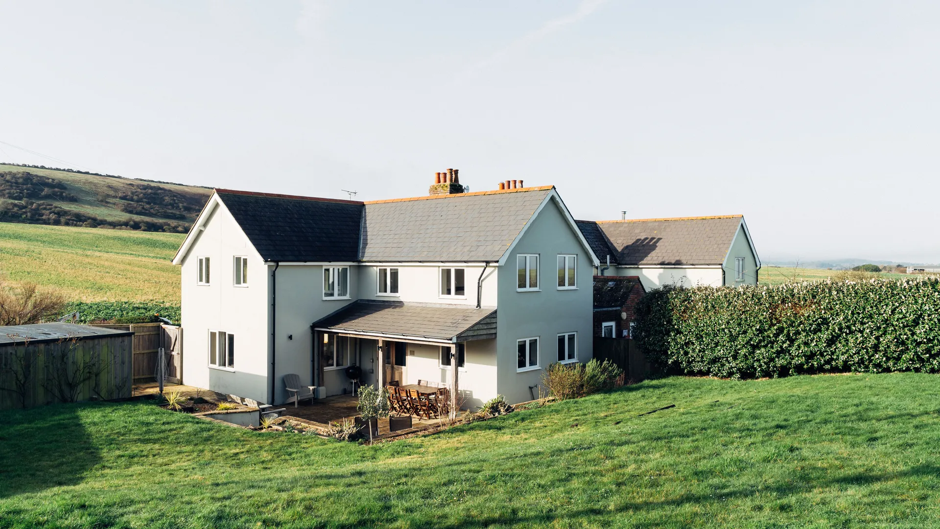 Modern countryside house with a patio and lawn, set against rolling green hills under a clear sky.