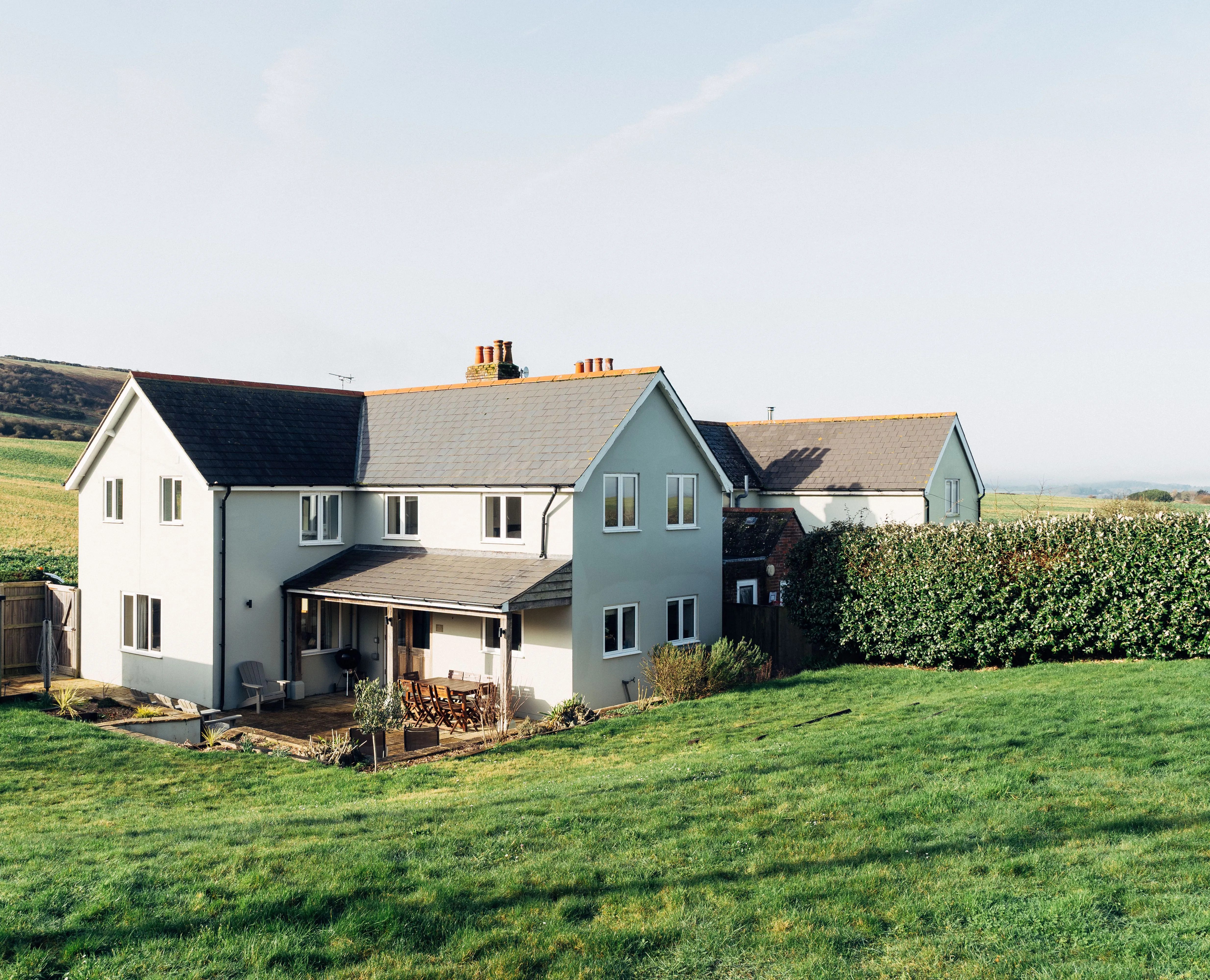 Modern countryside house with a patio and lawn, set against rolling green hills under a clear sky.
