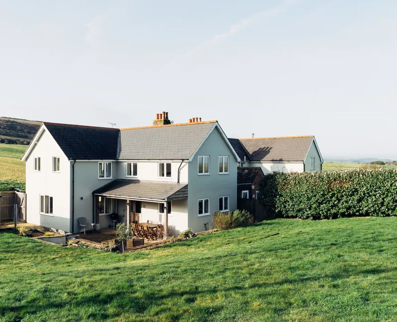 Modern countryside house with a patio and lawn, set against rolling green hills under a clear sky.