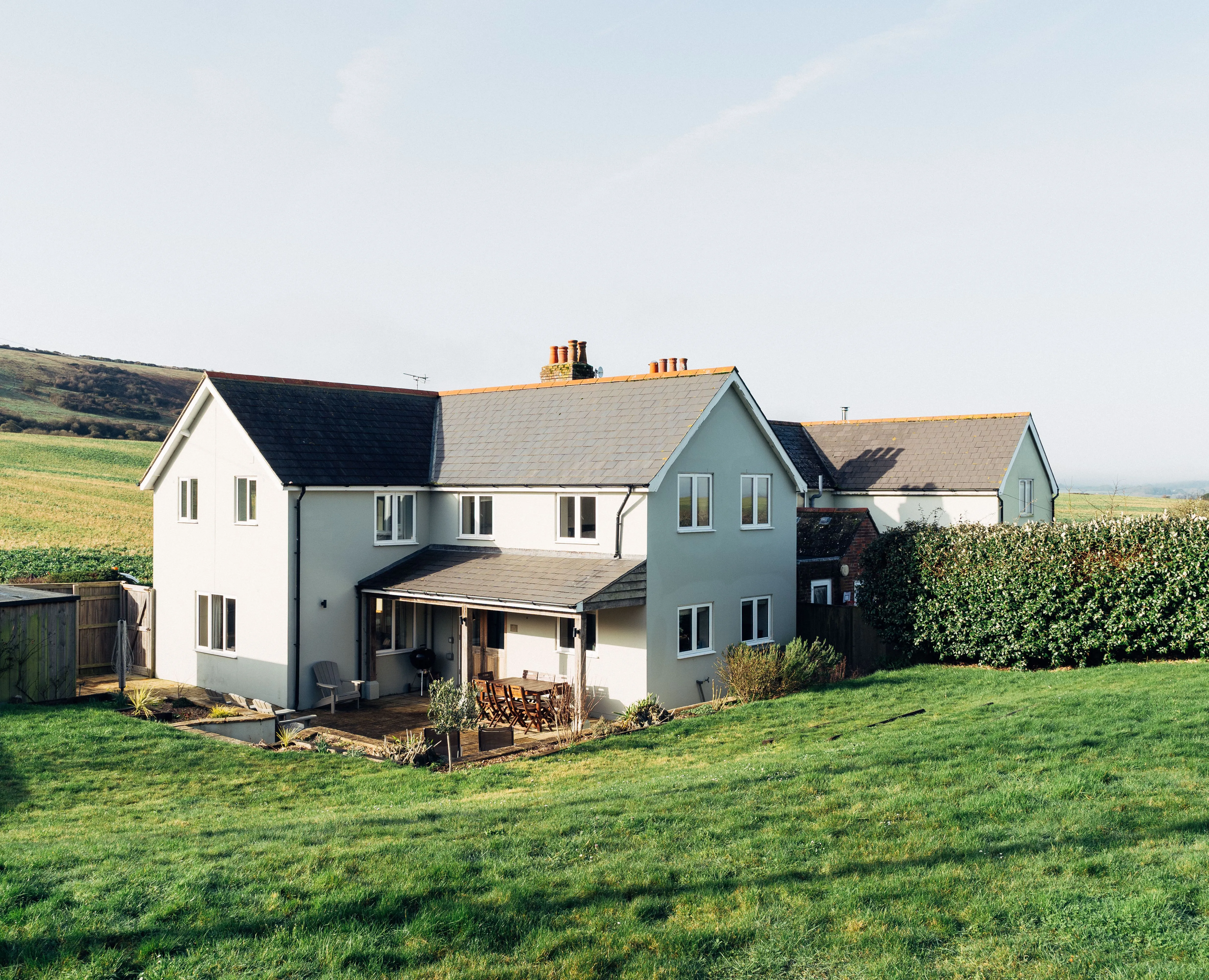 Modern countryside house with a patio and lawn, set against rolling green hills under a clear sky.