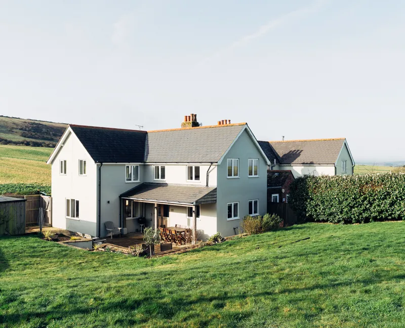 Modern countryside house with a patio and lawn, set against rolling green hills under a clear sky.