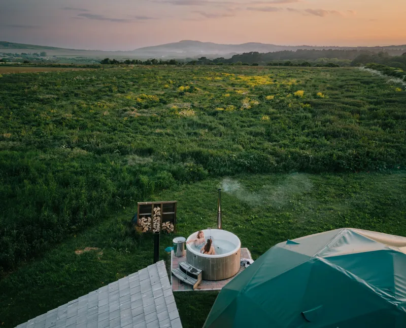 Aerial view of a countryside glamping site at sunset, with two people relaxing in a wood-fired hot tub beside a dome tent and open green fields.