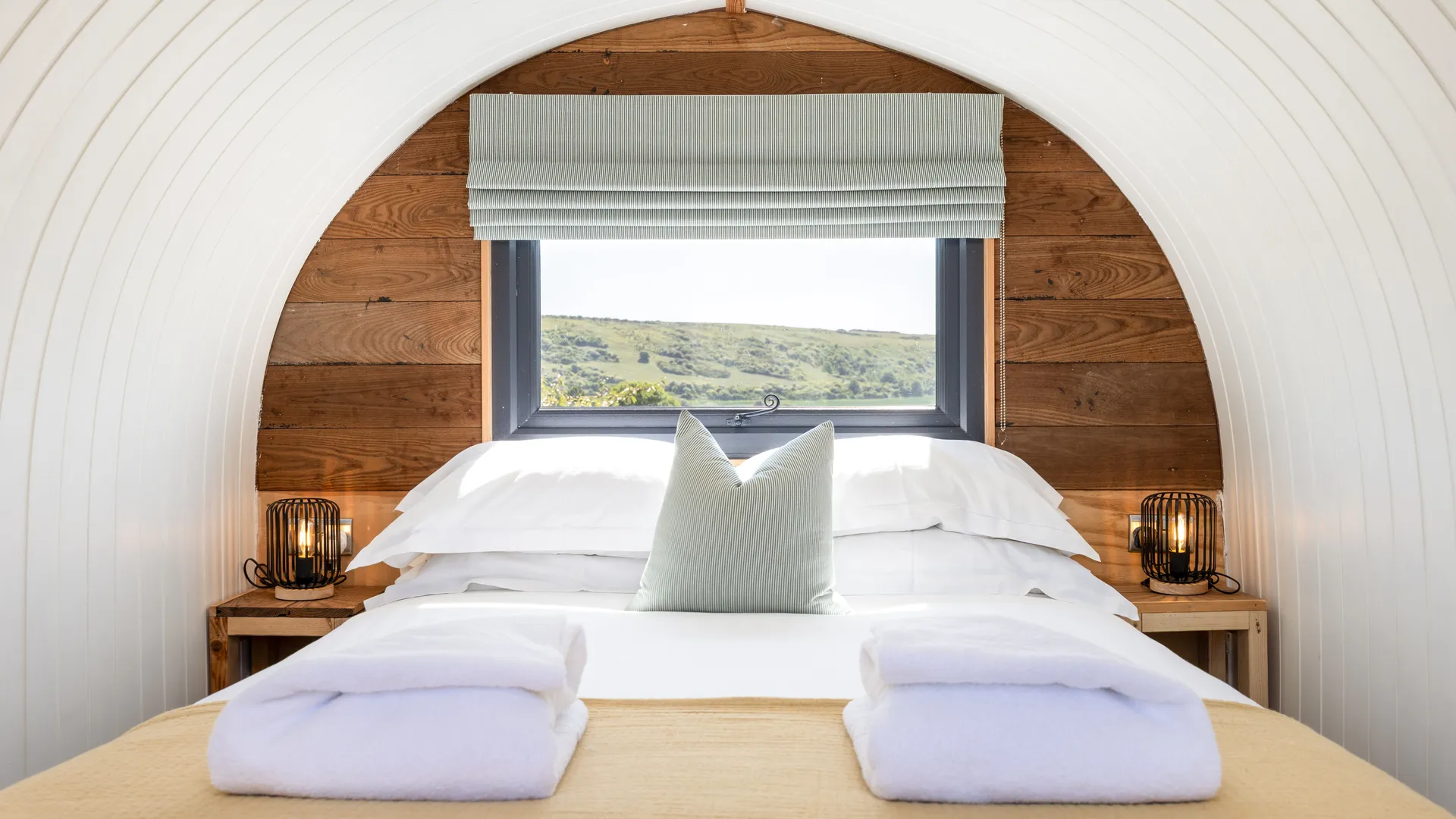 Cosy double bed inside a curved cabin with wooden panelling, bedside lamps and countryside views through the window