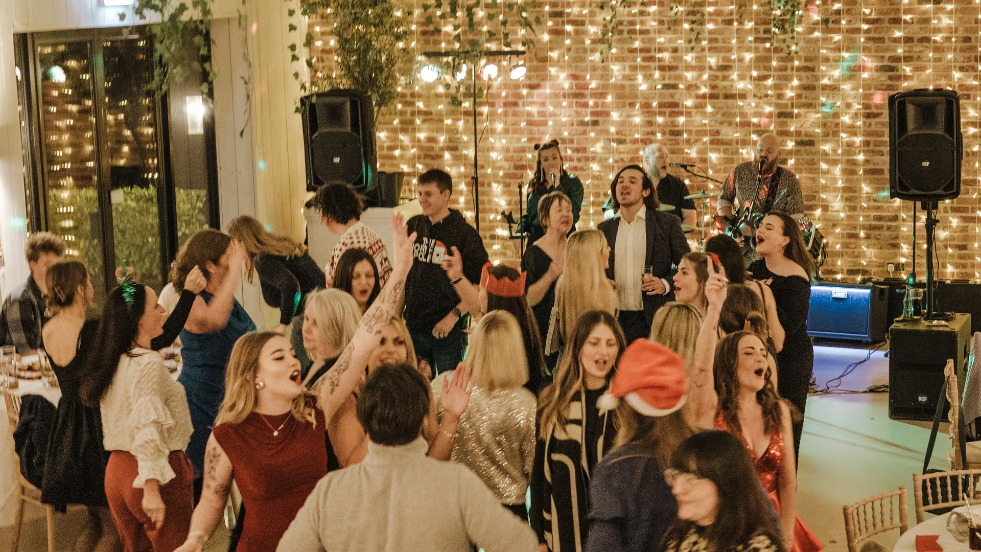 Guests dancing at a festive indoor party while a live band performs against a brick wall with fairy lights.
