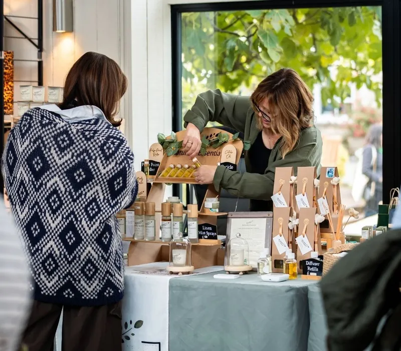 Woman arranging home fragrance products at an interiors show stall while a visitor browses.