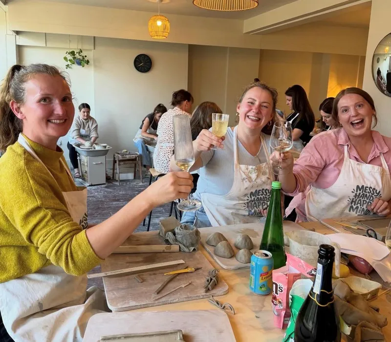 Three women in aprons raise glasses at a pottery workshop, seated around a table with clay, tools and drinks.