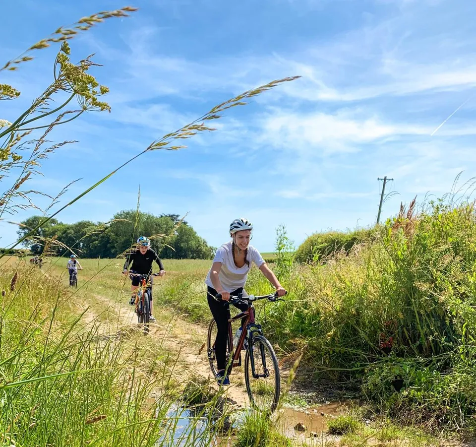 Cyclists riding along a grassy countryside trail with a shallow stream under a bright blue sky