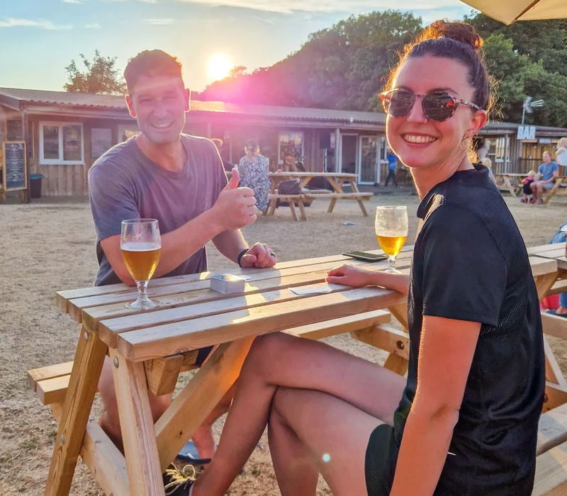 Two people enjoying drinks at a picnic bench during a live music evening at Tapnell Farm, with the sun setting behind them.