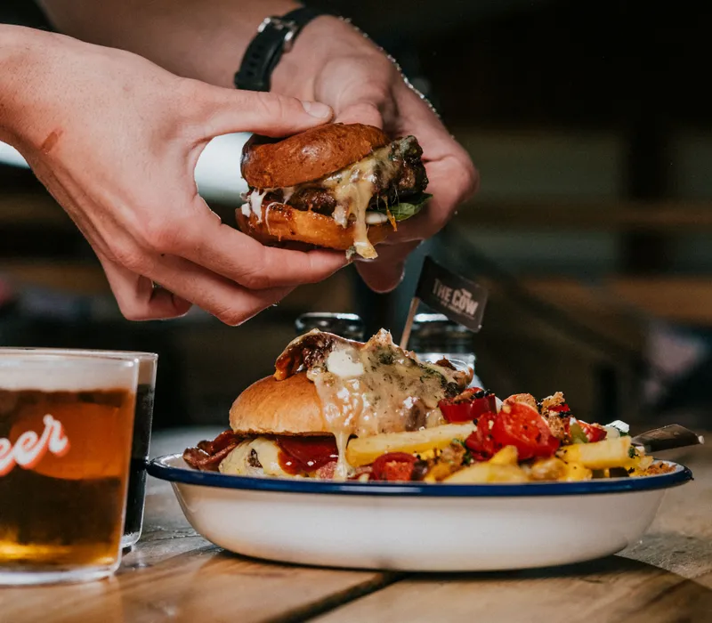 Hands holding a small cheeseburger above a plate with a loaded burger, chips and a glass of beer on a wooden table