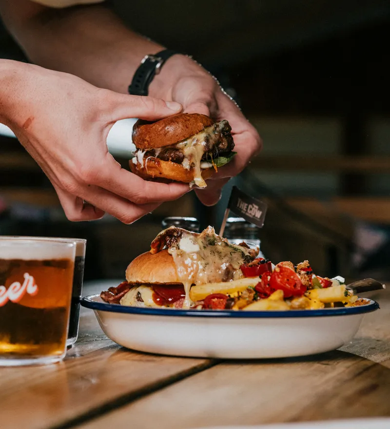 Hands holding a small cheeseburger above a plate with a loaded burger, chips and a glass of beer on a wooden table