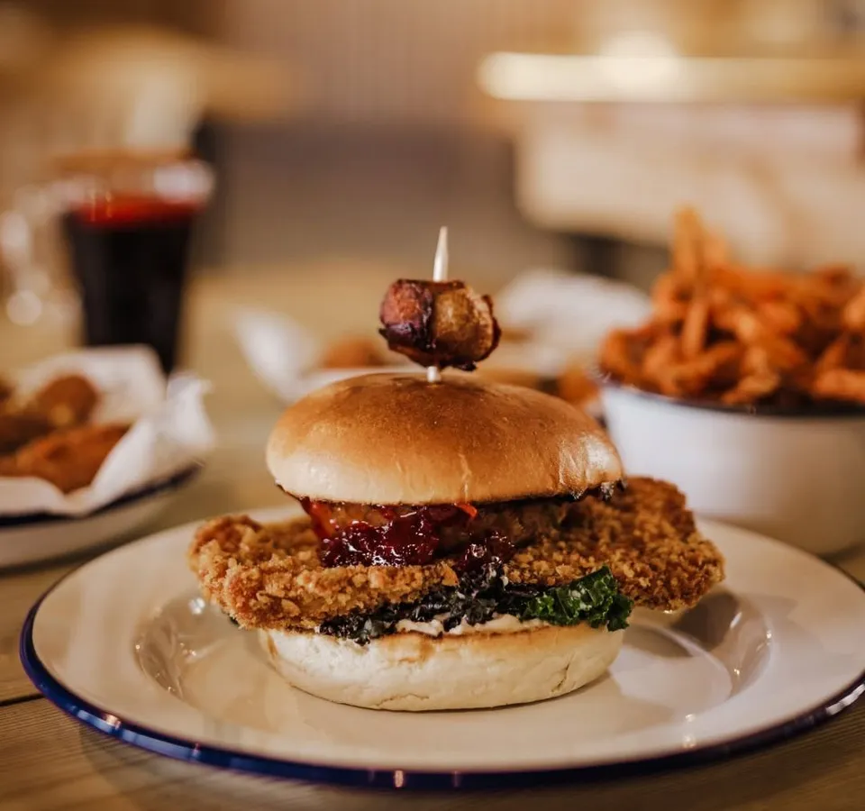 Festive crispy chicken burger with cranberry sauce, greens and a bacon garnish on a plate, served with chips and a soft drink in a warmly lit restaurant.