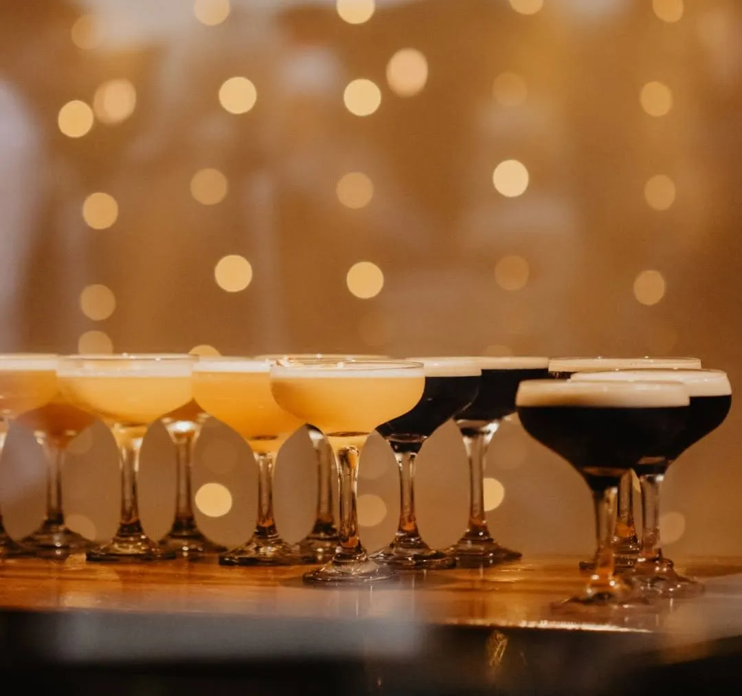 Cocktail glasses lined up on a bar with warm festive fairy lights in the background