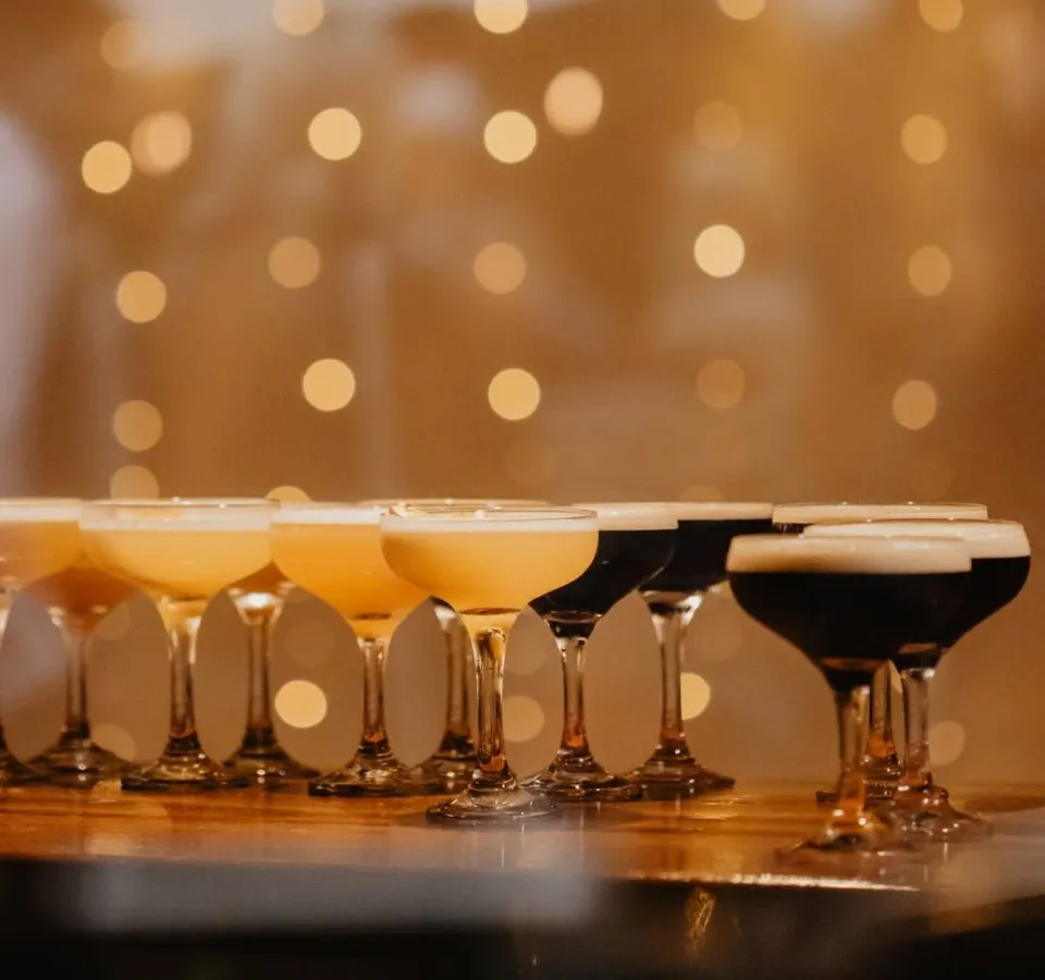 Cocktail glasses lined up on a bar with warm festive fairy lights in the background