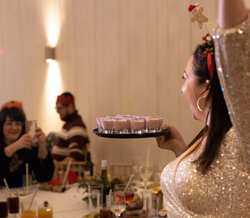 Woman in a sparkling gold dress carrying a tray of pink shots at a festive party