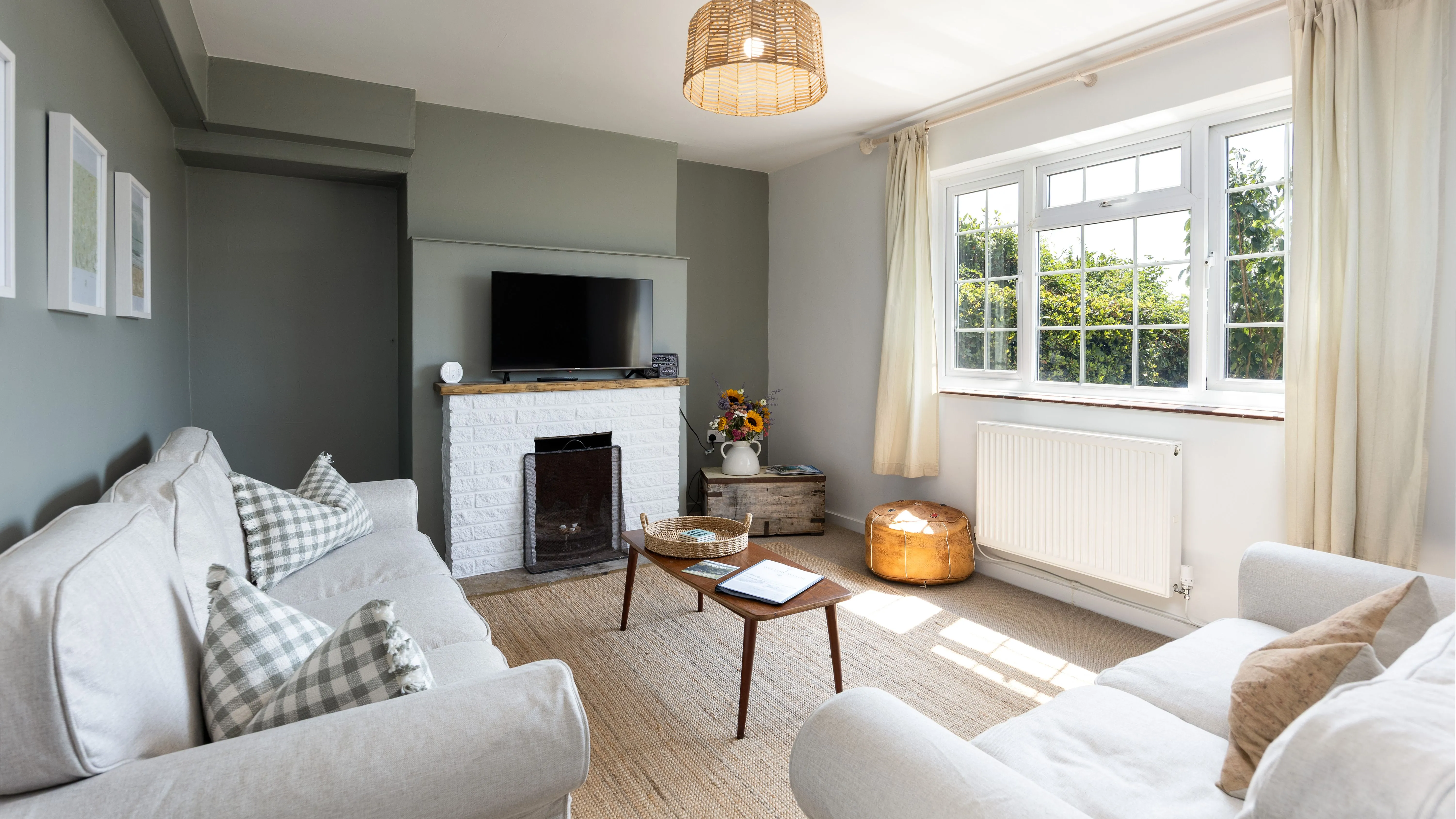 Bright, cosy cottage living room with pale sofas, a television above a white brick fireplace and large curtained windows.