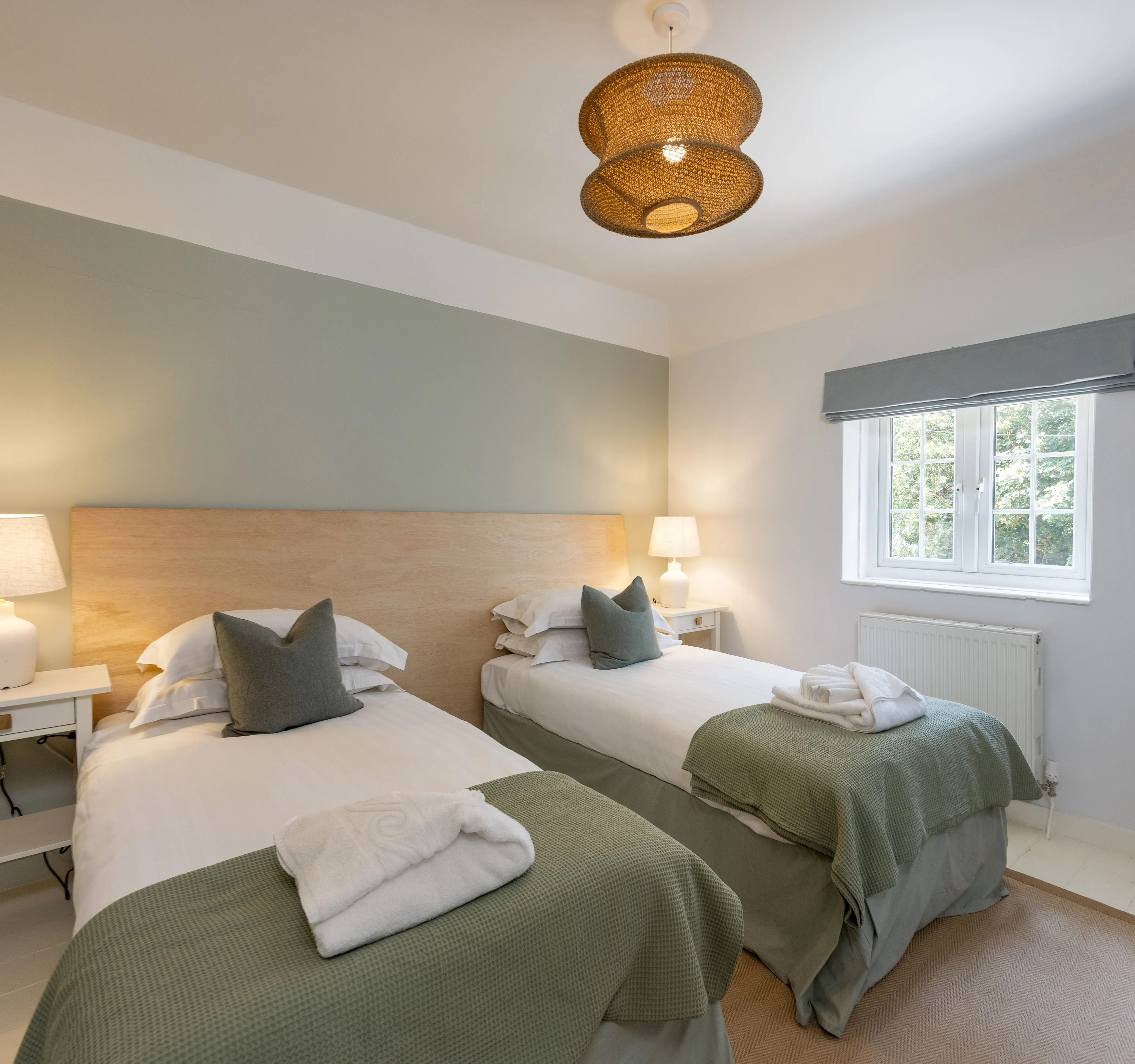 Cosy twin bedroom with sage green bedding, bedside lamps and a woven ceiling light.