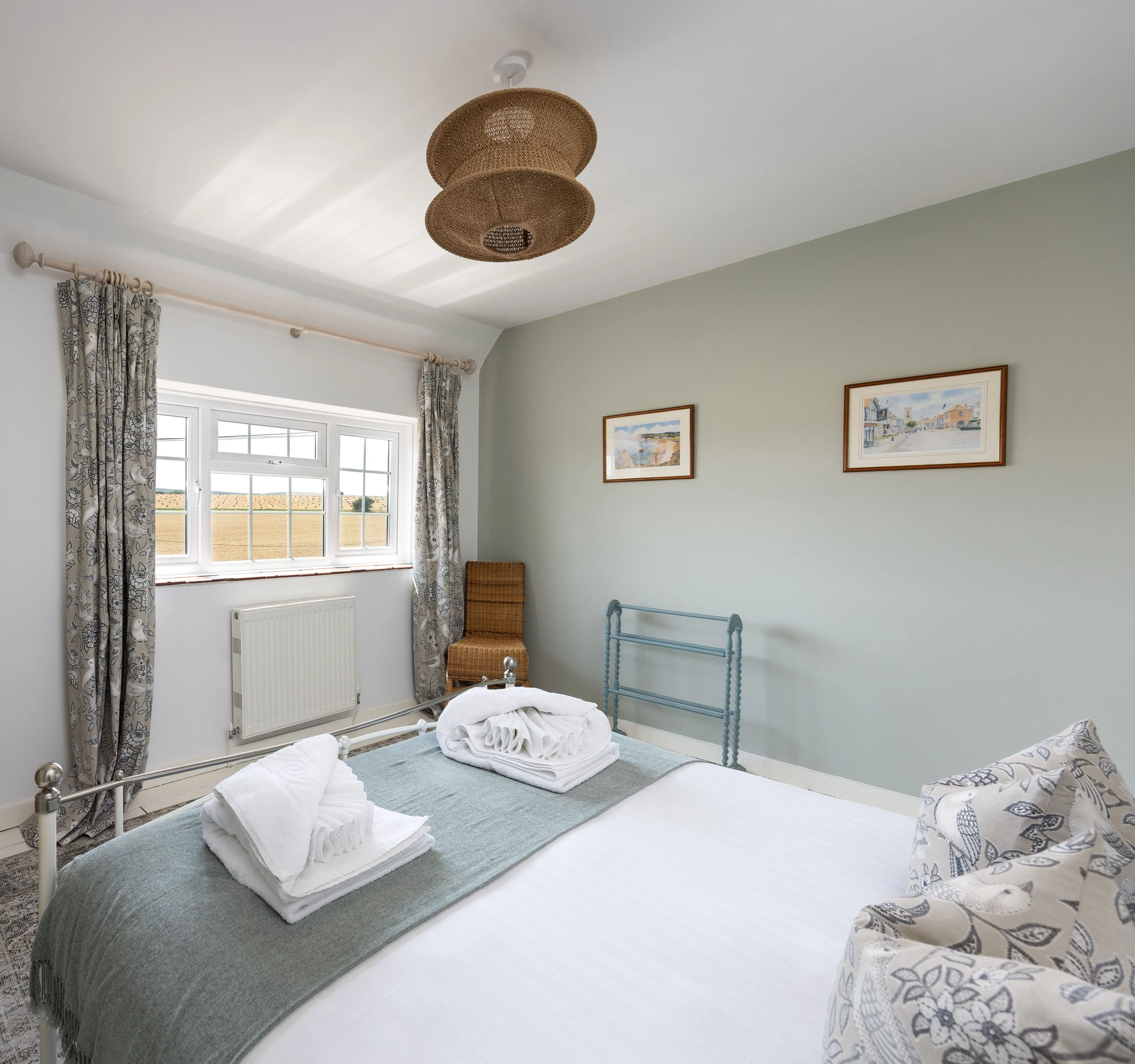 Bright cottage bedroom with a metal-framed double bed, folded towels, patterned curtains and rustic wooden furniture.