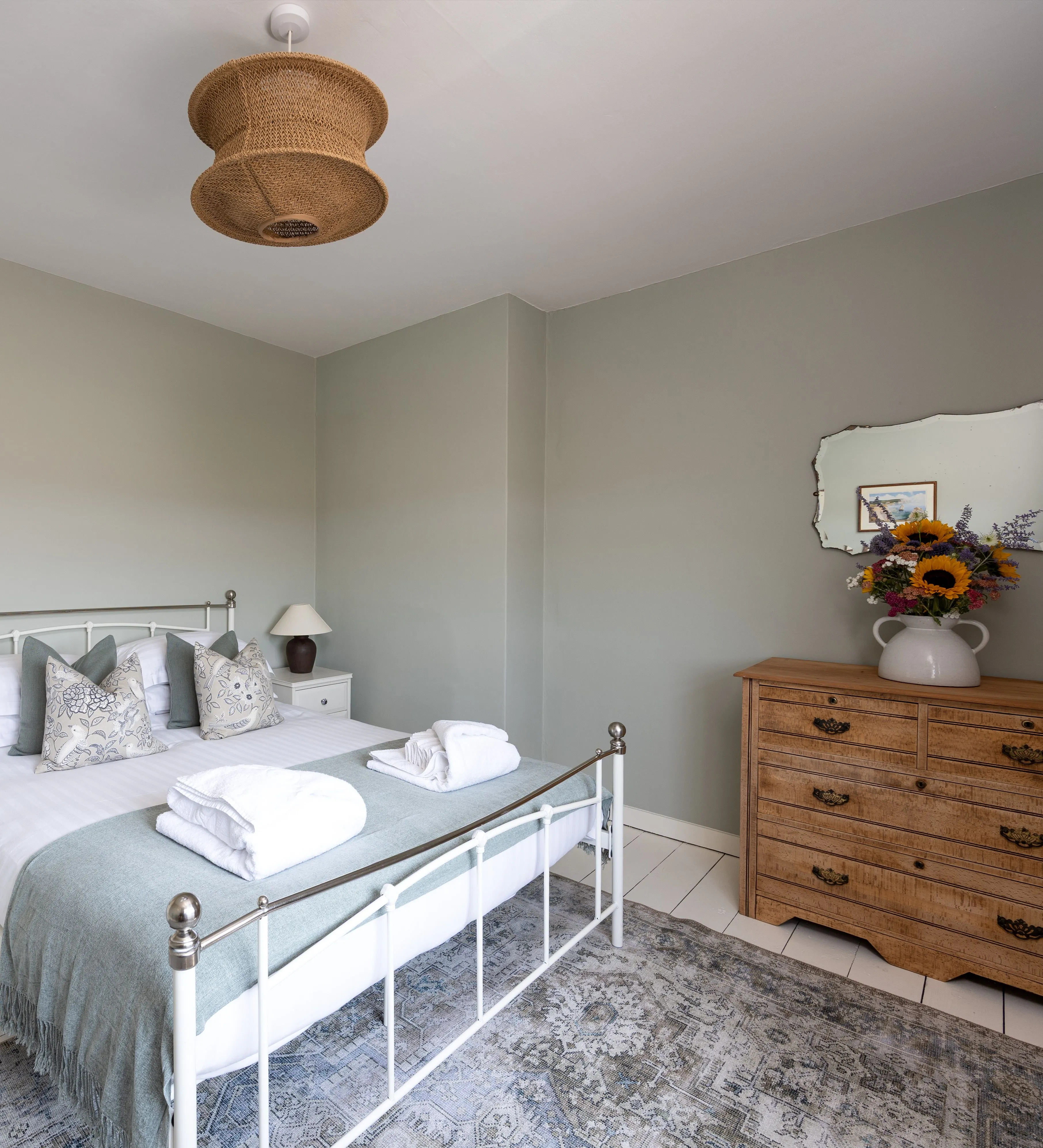 Bright cottage bedroom with a white metal bed, sage green walls and wooden chest of drawers.