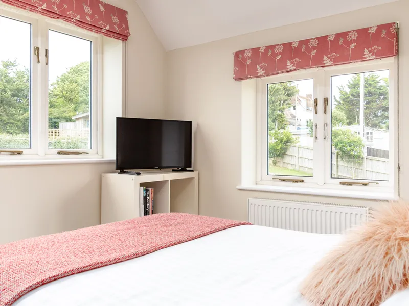 Bright bedroom with double bed, pink throw, television on a small unit and two windows with pink patterned blinds