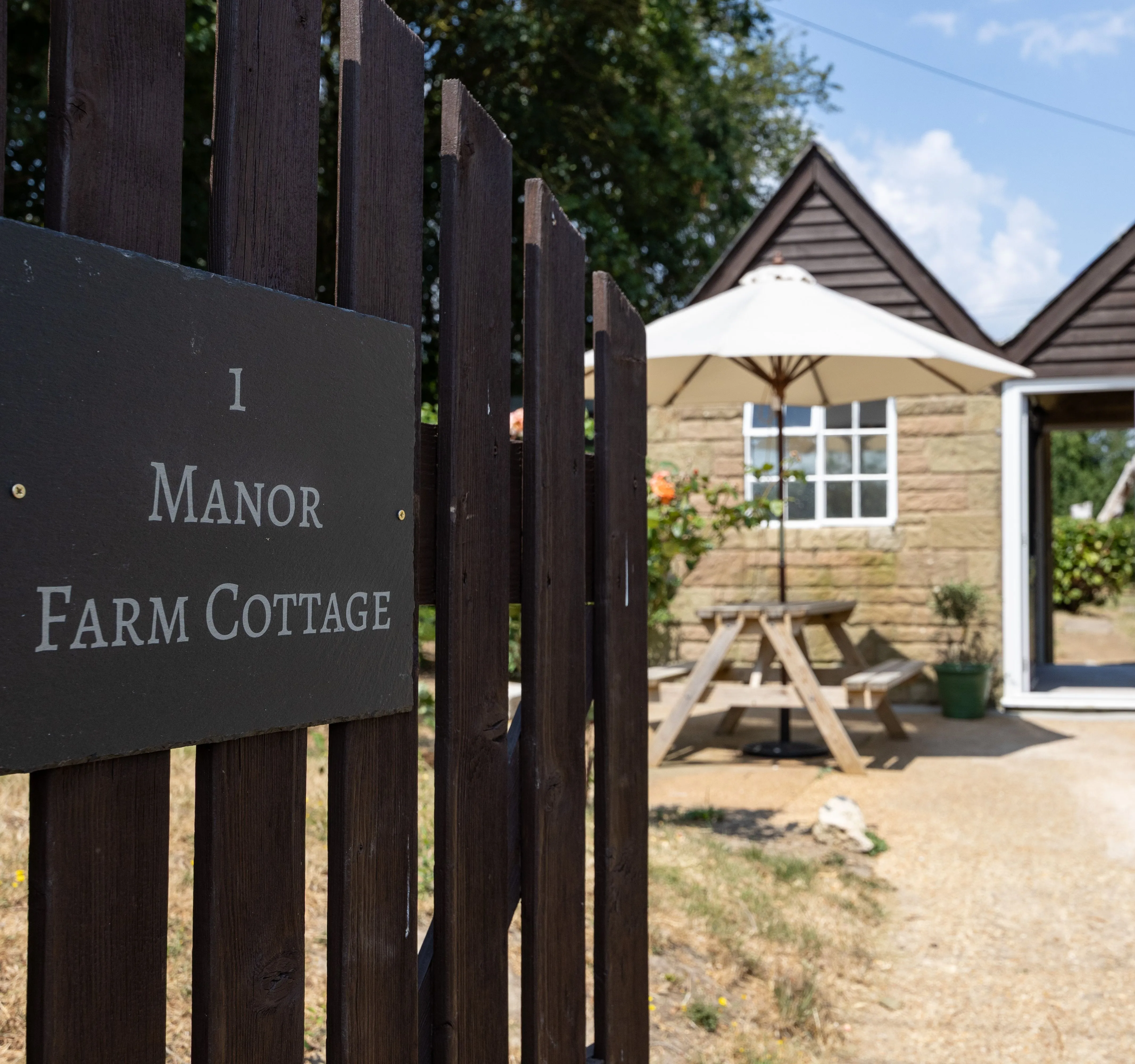 Entrance gate sign for Manor Farm Cottage beside a stone cottage patio with picnic table and parasol.