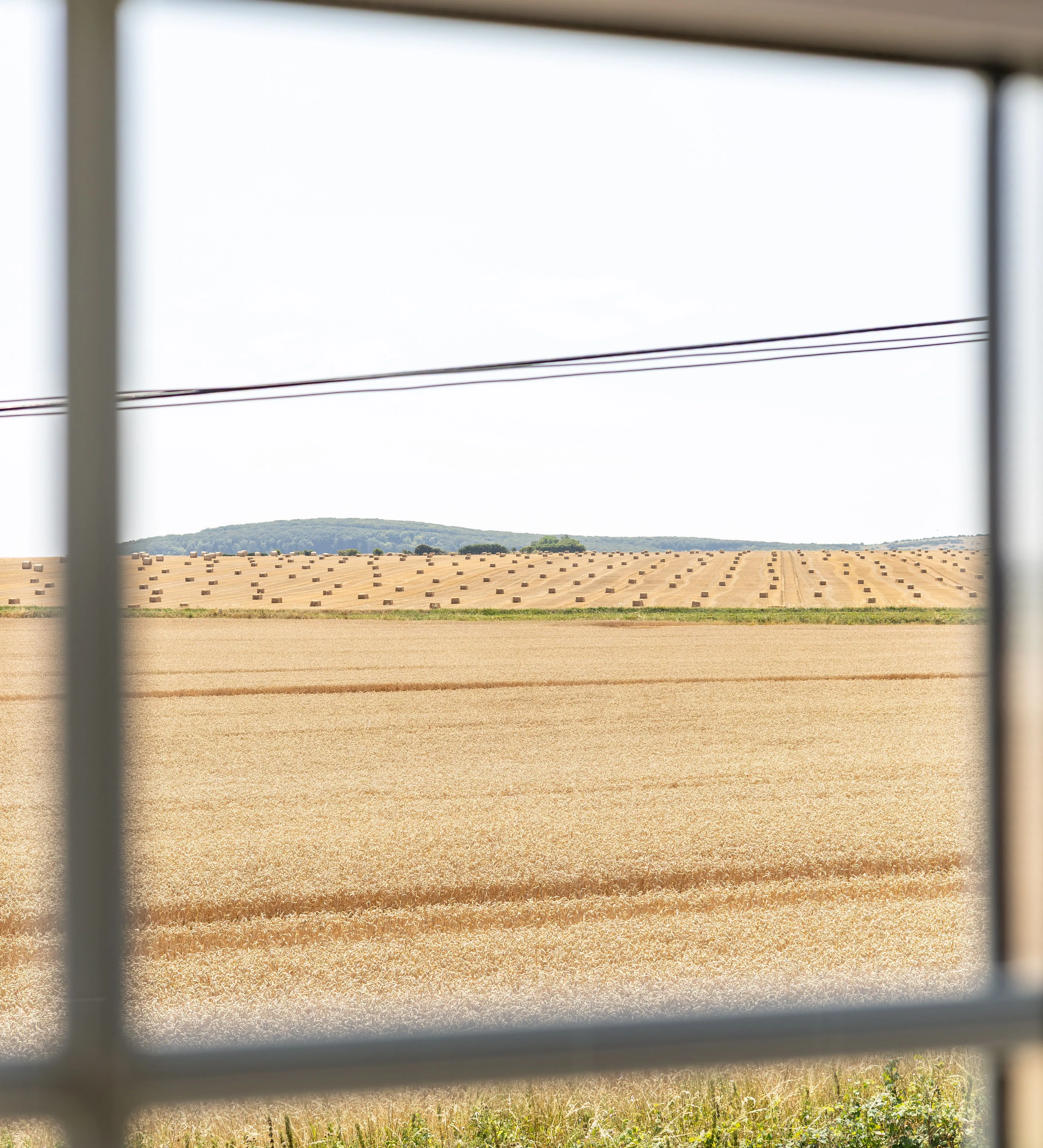 View through a cottage window across golden harvested fields dotted with hay bales.