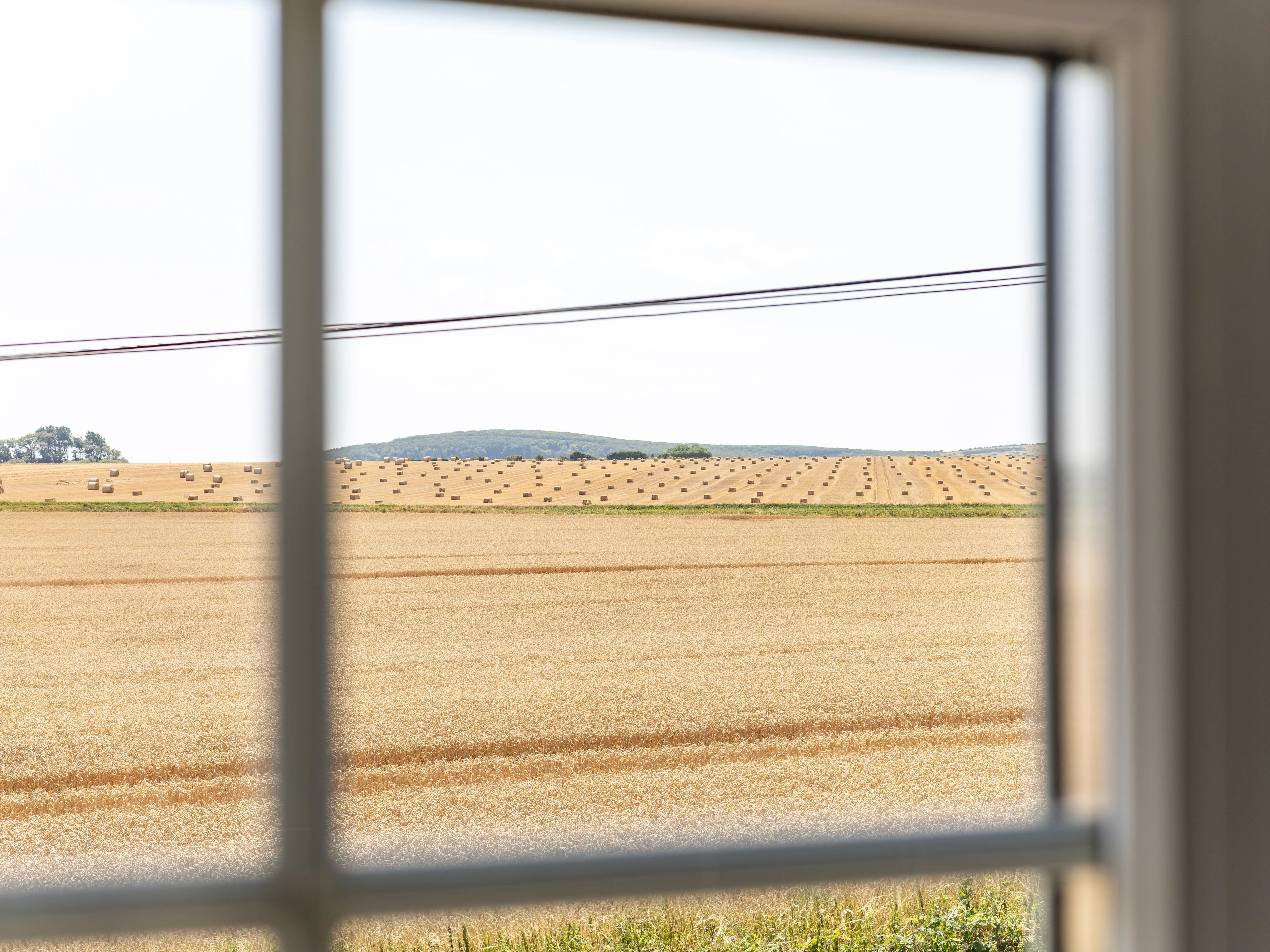 View through a cottage window across golden harvested fields dotted with hay bales.