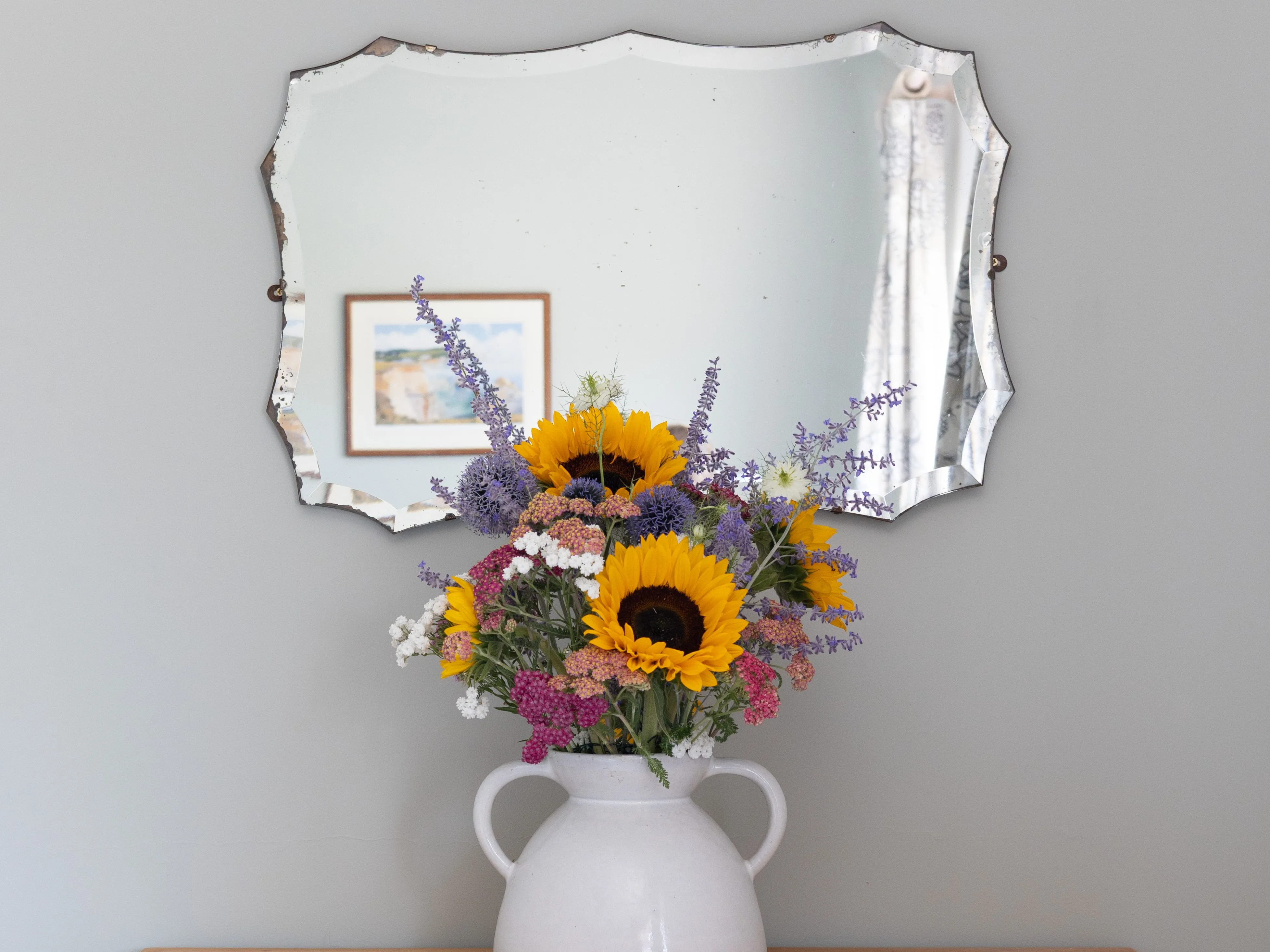 Colourful sunflower bouquet in a white ceramic jug on a vintage wooden chest beneath an ornate wall mirror.