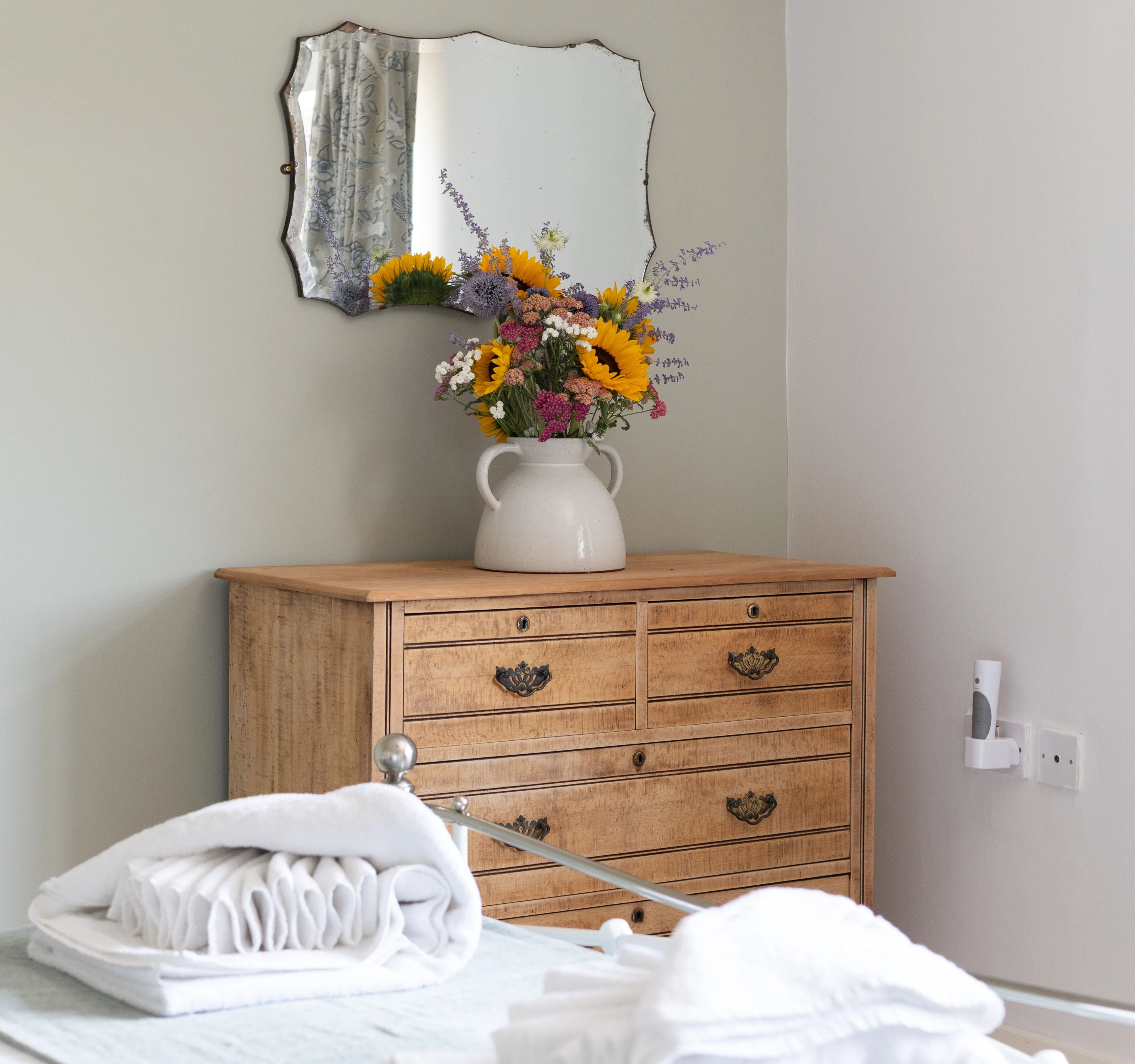 Cosy bedroom with folded white towels, a wooden chest of drawers and a vase of sunflowers beneath a vintage mirror.