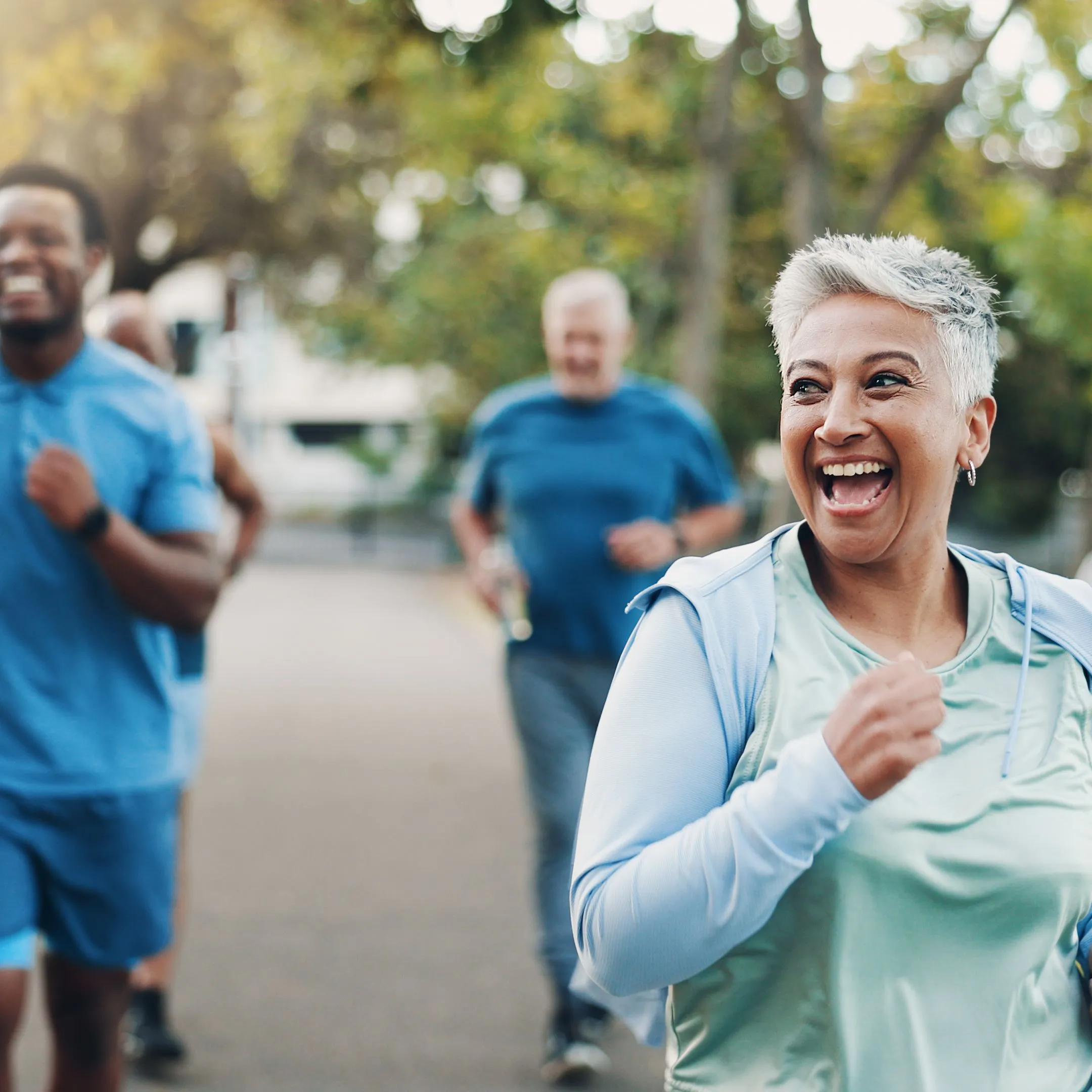 Smiling older woman jogging with a group of runners on a tree-lined park path.