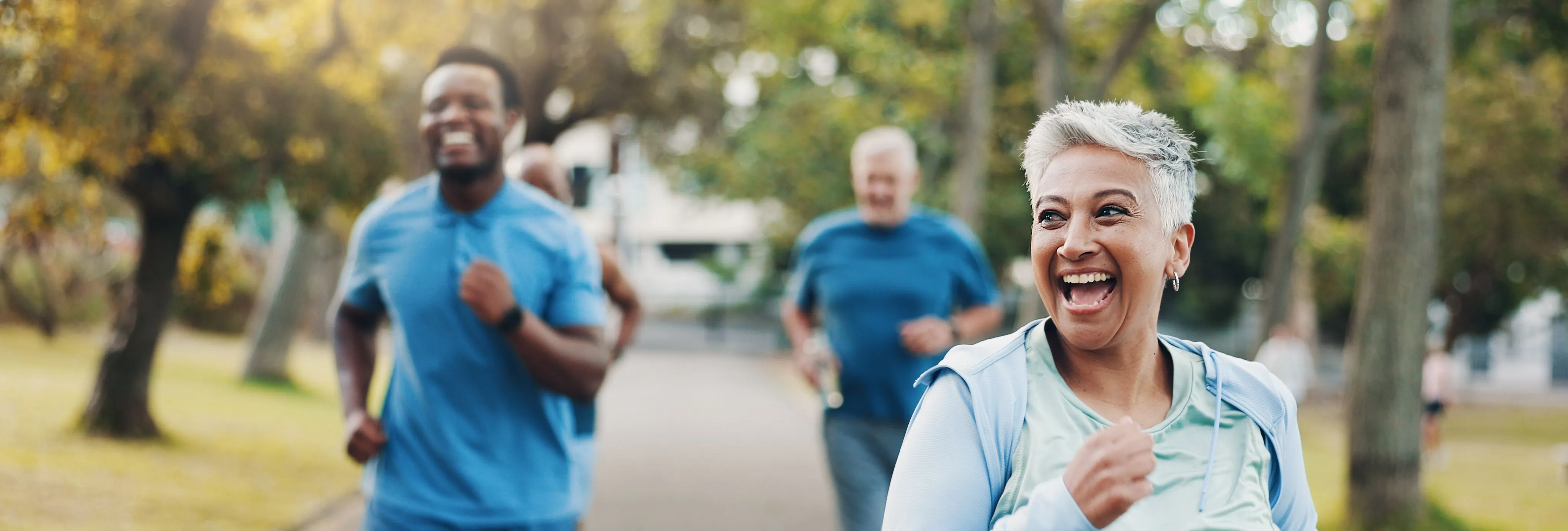 Smiling older woman jogging with a group of runners on a tree-lined park path.