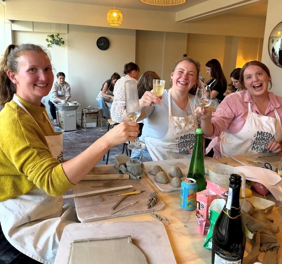 Three women in aprons raise glasses during a lively pottery workshop, surrounded by clay and sculpting tools.