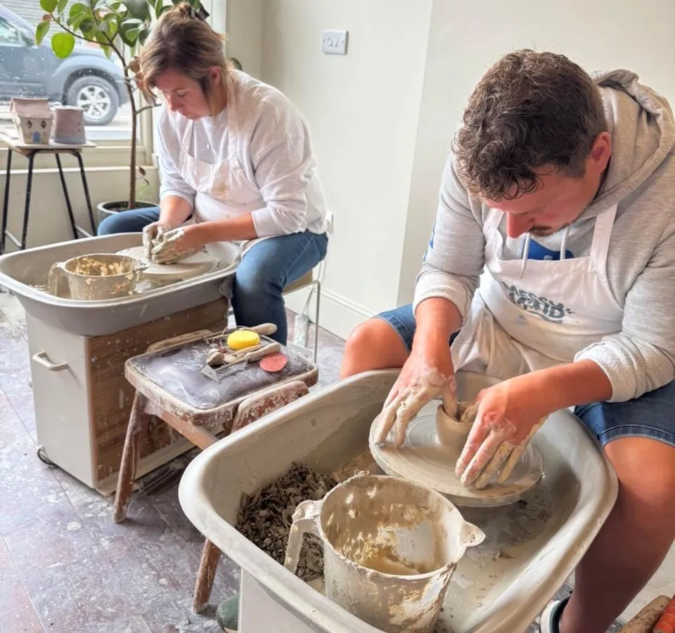 Two adults shaping clay on pottery wheels in a bright studio