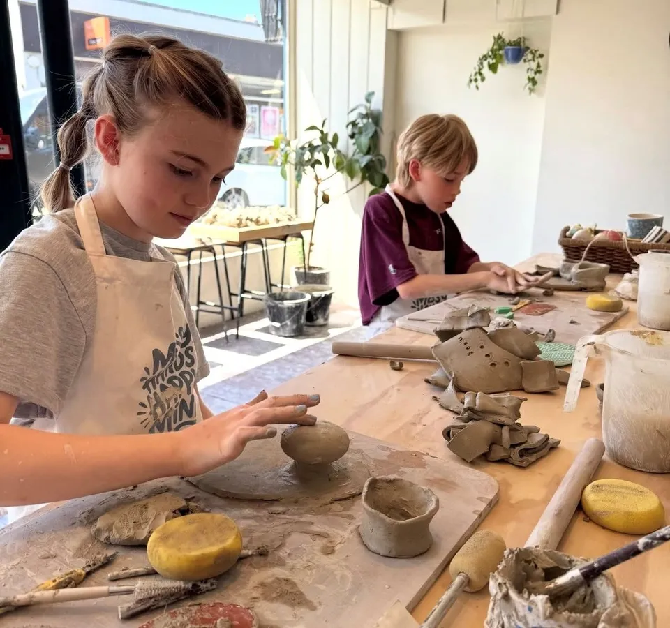 Two children wearing aprons shape clay at a table in a bright pottery studio.