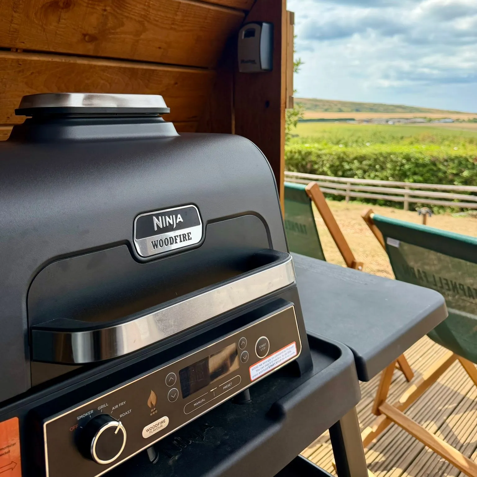 Ninja Woodfire outdoor grill on a wooden deck beneath a timber shelter, overlooking green countryside.