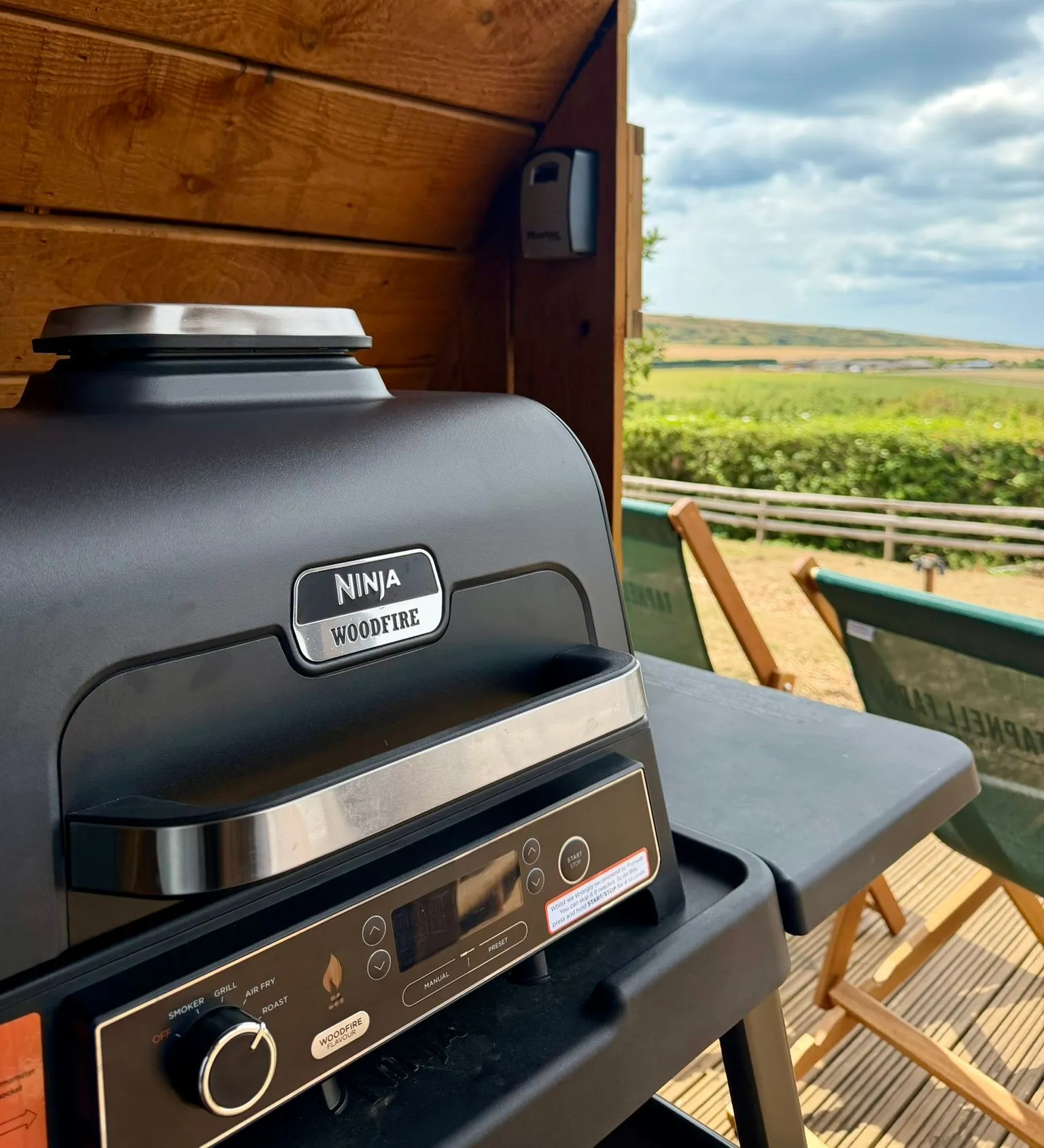 Ninja Woodfire outdoor grill on a wooden deck beneath a timber shelter, overlooking green countryside.