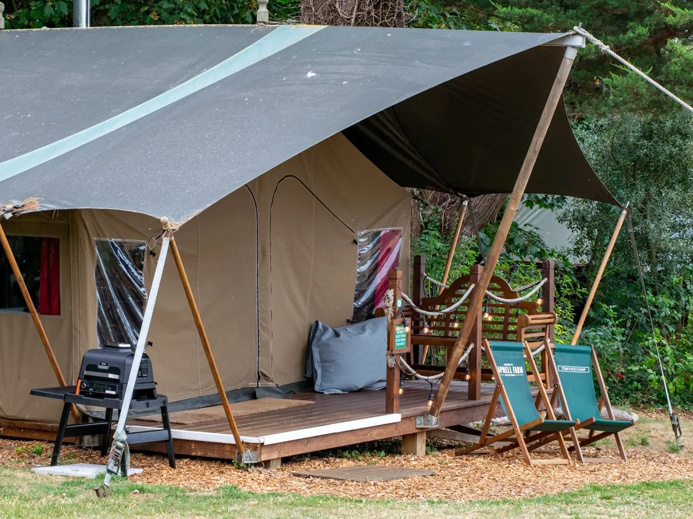 Canvas glamping tent with an electric barbecue and deck chairs in a wooded campsite.