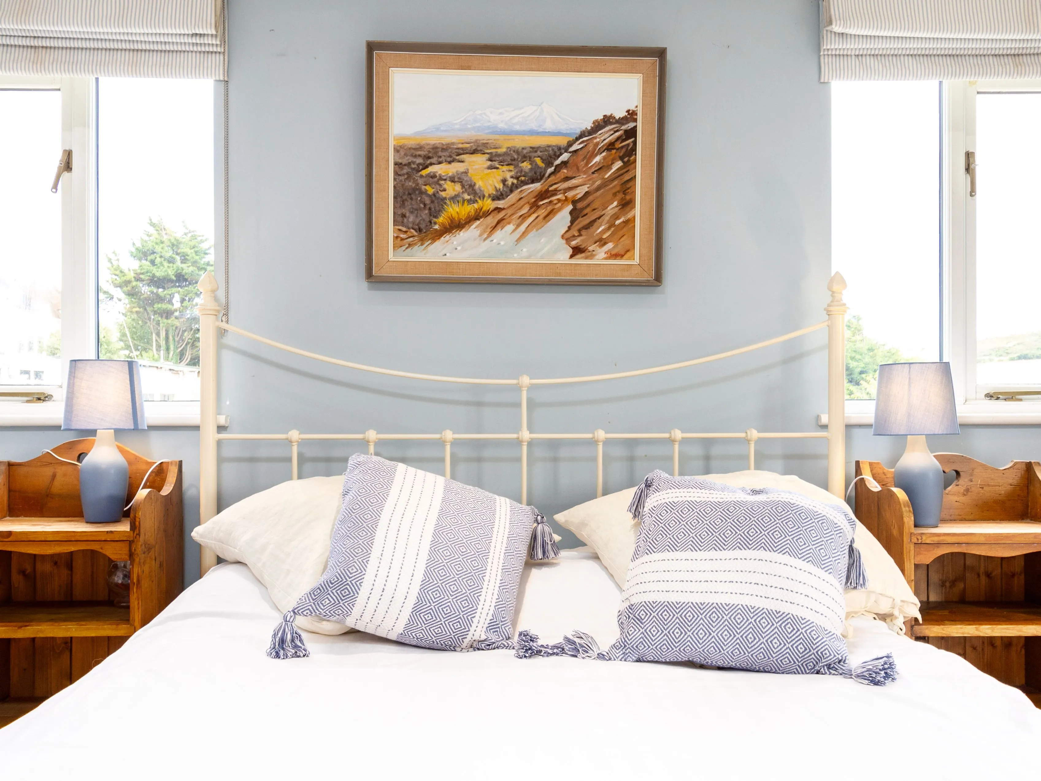 Bright bedroom with white metal bed, blue walls, patterned cushions and bedside lamps by two windows
