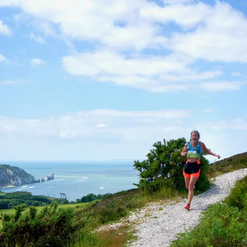 Female trail runner on a chalky coastal path overlooking the sea and cliffs under a blue sky.